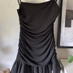 Svart kort klänning med rynkade detaljer - Säljer en svart kort klänning med smala axelband och rynkade detaljer både fram och bak. Klänningen har en volang nertill som ger en extra touch. Perfekt för dig som vill ha en stilren men ändå trendig look.💕