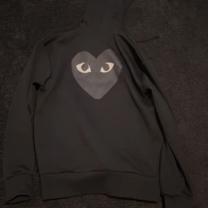Svart hoodie från Comme des Garçons Play - Svart hoodie med dragkedja och huva från Comme des Garçons Play. På bröstet finns det ikoniska hjärtat med ögon i grått. Perfekt för en stilren och cool look. Nypris 2500kr