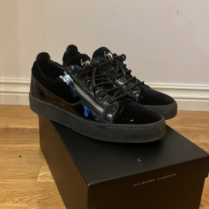 Svarta sneakers från Giuseppe Zanotti med dragkedjor - Säljer ett par svarta sneakers från Giuseppe Zanotti med snygga silvriga dragkedjor på sidorna och svarta snören. Skorna har en platt sula och är tillverkade i en mix av skinn och syntetmaterial med glansiga detaljer.