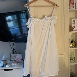 Vit axelbandsklänning med volang - Söt vit klänning med smala axelband och volangdetalj upptill. Klänningen har en luftig och avslappnad passform, perfekt för varma dagar. Materialet känns lätt och bekvämt mot huden.