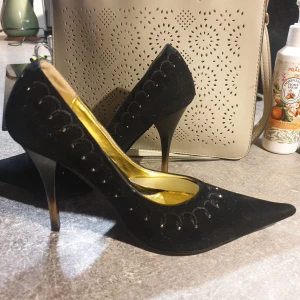 Svarta pumps med spetsig tå och klack - Säljer ett par eleganta svarta pumps i mocka med spetsig tå och hög klack. Skorna har dekorativa broderade detaljer längs ovandelen och guldfärgad innersula som ger en lyxig känsla. Perfekta för dig som vill sticka ut med stil.