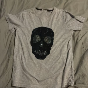 Grå t-shirt med paljett-dödskalle från H&M - Grå t-shirt från H&M med en stor svart paljett-dödskalle på framsidan. T-shirten har rund halsringning och är kortärmad. Perfekt för dig som gillar edgy detaljer och vill sticka ut lite extra.