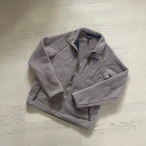 Ljusgrå fleecejacka från Patagonia - Mysig ljusgrå fleecejacka från Patagonia med hel dragkedja och tre fickor med dragkedja. Perfekt för kyliga dagar och enkel att matcha med olika outfits. Klassisk logga på bröstet och hög krage.