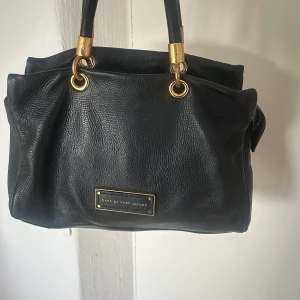 Svart handväska från Marc by Marc Jacobs - Säljer en klassisk svart handväska från Marc by Marc Jacobs med guldfärgade metalldetaljer och dubbla handtag. Väskan har en stilren design med logoplatta framtill och är tillverkad i läder