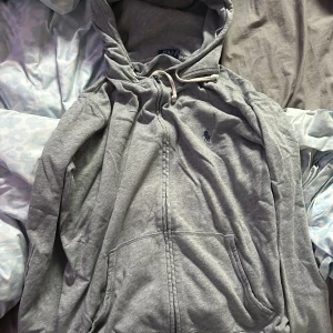 Grå hoodie med dragkedja från Polo Ralph Lauren - Säljer en grå hoodie från Polo Ralph Lauren med dragkedja och huva. Tröjan har fickor framtill och en liten broderad logga på bröstet. Perfekt för en avslappnad stil.