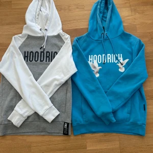  exklusiva special edition Hoodrich hoodies i grå/vit och blå - Säljer två snygga limited edition hoodies från Hoodrich. En är grå med vita ärmar och huva, svart tryck fram och text bak. Den andra är blå med vit tryck och duvor fram samt stort motiv bak. Båda har huva och känguruficka. Den blåa i storlek S och den gråa i storlek M