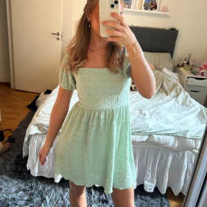 Ljusgrön kort klänning med puffärmar - Superfin ljusgrön klänning med smock upptill och kort puffärm. Klänningen har en söt, rynkad struktur och en utställd kjol. Perfekt för vår och sommar, och ger en fräsch look med sin pastelliga färg.