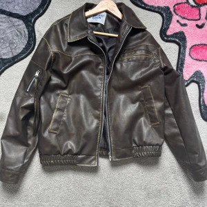 Brun skinnjacka från Elfric Eden - Snygg brun skinnjacka från Elfric Eden med dragkedja framtill, två snedställda fickor och en ficka med dragkedja på ärmen. Jackan har elastisk midja och klassisk krage. Perfekt för dig som vill ha en stilren och tidlös look.