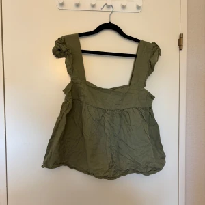Olivgrön volangtopp med breda axelband - Säljer en olivgrön topp med breda axelband och volangdetaljer. Toppen har en lös passform och är tillverkad i ett lätt tyg, perfekt för varmare dagar. Snygg att styla med jeans eller kjol!