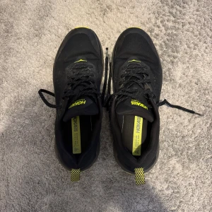 Svarta sneakers från Hoka One One med gula detaljer - Säljer ett par svarta sneakers från Hoka One One med gula detaljer och sula. Skorna har snörning och är tillverkade i syntetmaterial med meshpaneler för extra andningsförmåga. Perfekta för träning eller vardagsbruk.