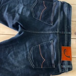 Mörkblå jeans från Jacob Cohën Style 622 - Säljer ett par mörkblå jeans från Jacob Cohën, modell Style 622. Jeansen har orangea kontrastsömmar, klassiska fem fickor och en snygg patch i orange på baksidan. Knapparna har märkets logga och passformen är normal.