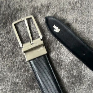 Svart perforerat skärp från Lacoste - Snyggt svart bälte från lacoste  skinn med perforerat mönster längs hela remmen. Klassisk silverfärgad metallspänne och den ikoniska krokodil logan i vitt vid spetsen. Perfekt accessoar för att lyfta din outfit med en sportig touch.