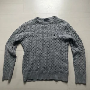 Grå kabelstickad tröja Ralph Lauren - Kabelstickad grå tröja från Ralph Lauren med rund halsringning och diskret broderad logga på bröstet. Långärmad modell i mjuk bomull, perfekt för lager-på-lager och chill stil. Den är i storlek M/S