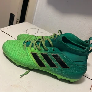 Gröna adidas fotbollsskor med snörning - Snygga gröna adidas fotbollsskor med svarta ränder på sidorna och högt skaft. Skorna har snörning och är tillverkade i syntetmaterial med texturerad ovandel för extra grepp. Perfekta för dig som vill sticka ut på planen.