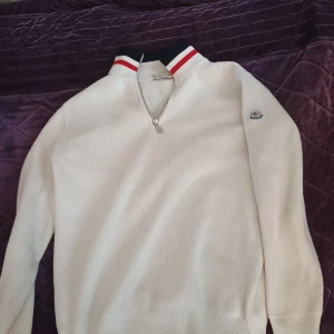 Vit stickad Moncler half zip tröja - Vit ribbstickad tröja från Moncler med halv dragkedja och hög krage med marinblå och röda ränder. Klassisk Moncler-logga på ärmen. Perfekt för dig som gillar stilren och sportig vibe.