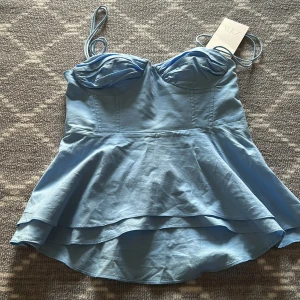 Ljusblå bustier-topp från Zara - Supersöt ljusblå bustier-topp från Zara med tunna axelband och volangig peplum-nederdel. Toppen har formade kupor och smockad rygg för extra komfort. Perfekt för att skapa en feminin och trendig look.