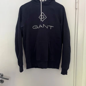 Mörkblå hoodie från GANT - Snygg mörkblå hoodie från GANT med vit logga och dragsko i huvan. Klassisk känguruficka framtill och mjukt material som känns skönt mot huden. Perfekt för dig som gillar en stilren och sportig look.