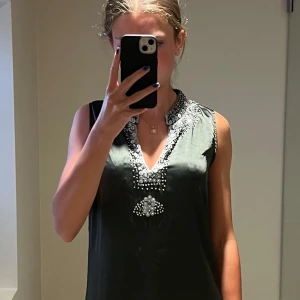 Svart kortklänning med paljetter från Indiska - Supersnygg svart kortklänning från Indiska i 100% polyester. Klänningen är ärmlös och har en v-ringad hals med silvriga paljettdetaljer runt halsringningen och framtill. Perfekt för dig som gillar att sticka ut med lite extra bling.