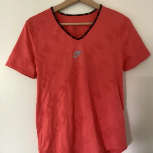 Nike Air korall t-shirt med öppen rygg Storlek S - Nike Air t-shirt i korallröd färg med diskret mönster och svart kant med AIR-tryck runt v-ringningen. Tillverkad i lätt polyester Dri-Fit material som andas. Cool detalj med öppen rygg för extra ventilation. Perfekt för dig som gillar sportig och trendig stil.