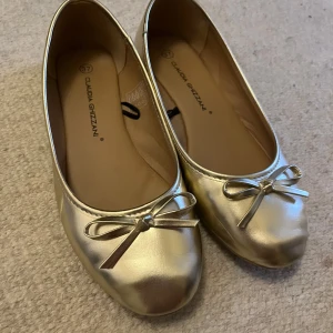 Guldiga ballerinaskor från Claudia Ghizzani - Snygga ballerinaskor i guldfärg! Skorna har rund tå, platt sula och en söt rosettdetalj framtill. Perfekta för dig som vill sticka ut med en glansig touch. 