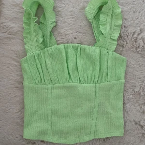 Ljusgrön linnetopp med volangband - Superfin ljusgrön linnetopp med breda axelband dekorerade med volanger. Toppen har en smockad struktur och är figurnära med en croppad passform. Perfekt för varma dagar när du vill sticka ut lite extra.