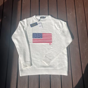 Vit stickad tröja Polo Ralph Lauren - Vit stickad tröja från Polo Ralph Lauren med amerikansk flagga broderad på bröstet och RL-detalj. Klassisk rund halsringning och ribbade muddar. Perfekt för dig som gillar preppy stil och vill sticka ut med en ikonisk detalj.
