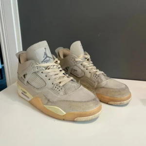 Nike Air Jordan 4 Off-White beige - Säljer ett par Nike Air Jordan 4 Off-White i beige med meshpaneler, detaljer i skinn och texten 'AIR' på sulan. Skorna har snörning, platt sula och ikonisk Jumpman-logga på plösen. Perfekta för dig som gillar streetwear och unika sneakers.