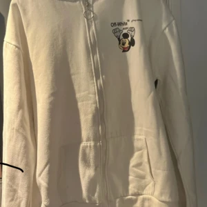 Vit Off-White hoodie med Mickey Mouse - Säljer en vit hoodie från Off-White med dragkedja och huva. Framsidan har ett litet tryck av Mickey Mouse och Off-White-logga, medan ryggen har en stor grafisk print med Mickey Mouse och Off-Whites ikoniska pilar. Mjuk bomullskänsla och känguruficka framtill.
