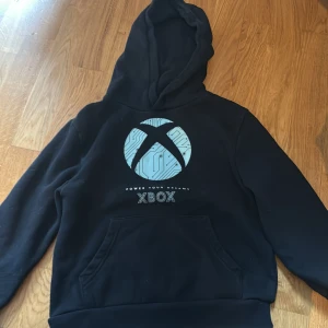 Svart Xbox hoodie med blå logga - Svart hoodie från Xbox med stor blå logga och texten 'Power Your Dreams' på bröstet. Tröjan har huva, magficka och långa ärmar. Perfekt för gaming eller chill. Mjuk insida och klassisk passform.