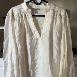 Ljusbeige v-ringad blus - Luftig och stilren ljusbeige henley blus med v-ringning och långa ärmar. Blusen har en avslappnad passform och är tillverkad i ett lätt material som passar perfekt för varmare dagar. Snygga rynkade detaljer vid axlarna ger extra karaktär.