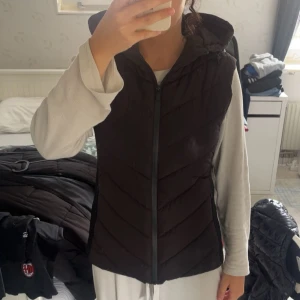 Svart dunväst från Zara med huva - Svart dunväst från Zara i storlek L. Västen har en quiltad design med snygga diagonala sömmar, huva och hel dragkedja framtill. Perfekt att slänga över en hoodie för extra värme. Materialet är polyester och västen har en normal passform.