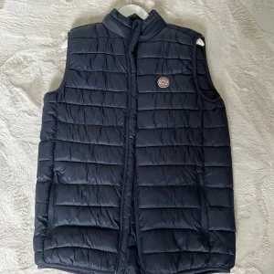 Mörkblå dunväst från Twentyfour - Snygg mörkblå  dunväst från kronstadt med quiltad design och hög krage. Västen har dragkedja framtill, två fickor med dragkedja och en diskret logga på bröstet. Perfekt att slänga över en hoodie för extra värme under kyliga dagar.