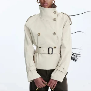 Beige trenchcoat med bälte och hög krage - Snygg beige trenchcoat med dubbelknäppning, hög krage och bälte i midjan. Jackan har långa ärmar med spännen vid mudden och markerad siluett. Perfekt för dig som gillar klassisk stil med modern twist. Från h&m med prislapp. Helt slutsåld. Köpt för 499