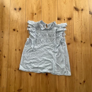 Blå blus med volang från Lindex - Supersöt blå blus från Lindex med volangdetaljer och broderade spetsinslag. Hög krage med liten volang och ärmlös design. Perfekt för dig som gillar romantisk och feminin stil.