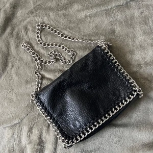 Svart axelväska med kedjedetaljer Tiamo - säljer en cool svart axelväska från Tiamo! väskan har dragkedja både på insidan och utsidan samt ett snyggt flätat mönster längs kanten. defekter på baksidan av väskan, därav priset. skriv vid fler frågor!💓