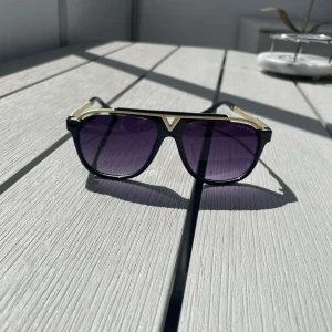 svarta och guldiga solglasögon - Snygga solglasögon från  med svart båge och guldiga detaljer. Glasen är mörklila och har en klassisk pilotform. Sidorna är prydda med Louis Vuitton-loggan och bågarna har en lyxig känsla med metallinslag.