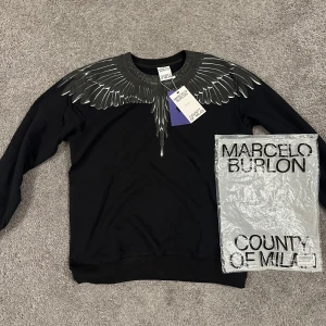 Svart tröja Marcelo Burlon vingar - Svart långärmad tröja från Marcelo Burlon County of Milan med ikoniskt vingtryck i grått och vitt över axlar och halsringning. Tröjan har rund hals och är gjord i mjuk bomull. Perfekt statement-piece för dig som gillar streetwear.