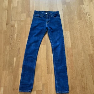 Blå skinny jeans från Nudie Jeans - Säljer ett par klassiska blå skinny jeans från Nudie Jeans i storlek W27 L32. Jeansen har fem fickor, snygga kontrastsömmar och Nudie-logga på bakfickan. Perfekta för dig som gillar en smal passform och tidlös stil.