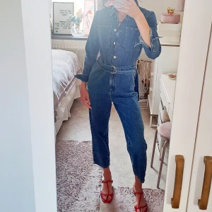 Blå jeans jumpsuit med bälte - Snygg blå jeans jumpsuit från NAKD, med croppade ben och tillhörande bälte i midjan. Modellen har raka ben, stora bröstfickor och knappar framtill. Materialet är klassisk denim och jumpsuiten har en cool, avslappnad vibe som passar perfekt för dig som gillar streetstyle. Nypris 900kr. 
