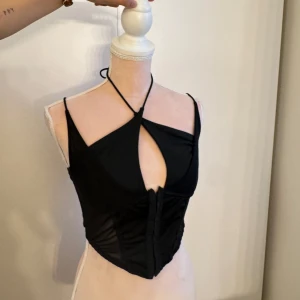 Svart korsett-topp med snörning - Trendig svart korsett-topp med smala axelband och halterneck-knytning. Toppen har en snygg cut-out framtill och markerad midja som ger en cool siluett. Perfekt för att lyfta vilken outfit som helst med en edgy vibe.