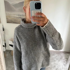 Grå stickad polotröja i loose fit - Mysig gråmelerad stickad polotröja med hög krage och avslappnad, loose passform. Tröjan har ett diskret mönster och långa ärmar. Stoleken är L men skulle säga att den är mer än S-M