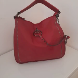 Röd axelväska med nitar och kedjedetaljer - Cool röd axelväska i skinn med silverfärgade nitar längs kanterna och chunky kedjedetaljer. Väskan har bred axelrem, dragkedja upptill och flera innerfack. Perfekt för dig som vill sticka ut med en edgy accessoar. Snabb leverans, priset kan diskuteras