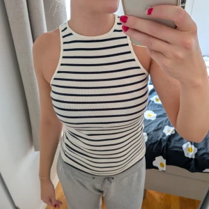 Randig ribbad linnetopp i vitt och svart - Säljer en snygg ribbad linnetopp med svarta och vita horisontella ränder. Toppen har hög halsringning och är ärmlös, perfekt för varma dagar. Den är figurnära och stretchig, vilket gör den både bekväm och trendig.