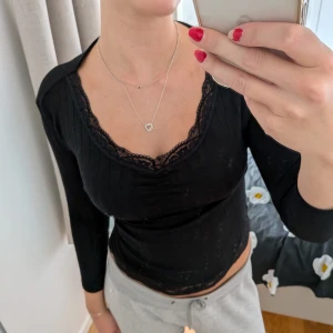 Svart ribbad långärmad topp med spets - Svart långärmad topp med ribbad struktur och v-ringning. Spetsdetaljer längs halsringning och nederkant ger en extra snygg touch. Sitter tight och har en feminin, croppad passform. Perfekt för dig som gillar stilrena men ändå detaljerade plagg.