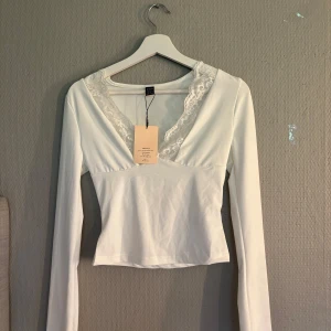 Vit långärmad topp med spets från SHEIN - Supersöt vit långärmad topp från SHEIN med djup v-ringning och fina spetsdetaljer längs halsen. Sitter tajt och har en clean, enkel look som passar perfekt till jeans eller kjol. Materialet är mjukt och stretchigt för en bekväm känsla.