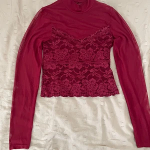 Vinröd meshblus med spetsdetaljer - Vinröd långärmad meshblus med hög hals och vackra blommiga spetsdetaljer över hela framsidan. Blusen är genomskinlig i ärmarna och har en feminin, romantisk look med fina kanter nertill. Perfekt att styla med jeans eller kjol.