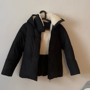 Svart pufferjacka från McKinley - Svart pufferjacka från McKinley med mysigt vitt foder och huva. Jackan har dragkedja framtill och en enkel, stilren design. Perfekt för kalla dagar när du vill hålla dig varm och bekväm. Passar dig som gillar en klassisk look med lite extra fluff.