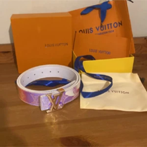 Louis Vuitton holografiskt bälte - Säljer ett Louis Vuitton bälte med holografisk yta i blått, rosa och lila toner. Bältet har den klassiska LV-loggan i metall som spänne och präglat monogrammönster längs hela remmen. Perfekt statement piece för att lyfta vilken outfit som helst.