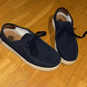 Mörkblå mockaloafers med snörning - Snygga mörkblå loafers i mocka med klassisk snörning och vit sula. Skorna har en rund tå och stilren design som passar perfekt till jeans eller chinos. Bekväm innersula och diskreta sömmar ger en clean look. Orginal låda medföljs ej. 