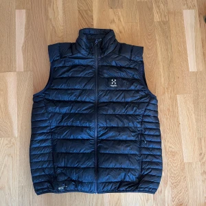  Haglöfs - Snygg svart dunväst från Haglöfs med quiltad design och hög krage. Västen har dragkedja framtill och diskret logga på bröstet. Perfekt för lager-på-lager och outdoor-vibes. Lätt och smidig att bära, passar till många olika outfits.